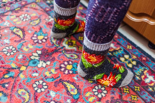 legs in warm woven wool socks standing on red ornate carpet floor at home backlit with a window light