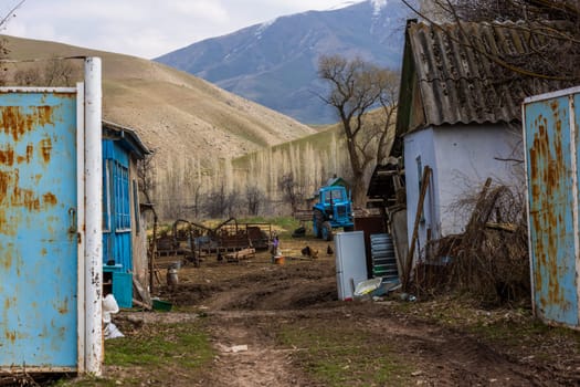 Poor rustic backyard with old blue tractor with dirt road between houses behind rusted steel gate at sunny spring day