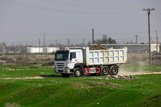 white Chinese dump truck loaded with pile of dirt and round rocks driving along dirt road at industrial area at sunny summer day