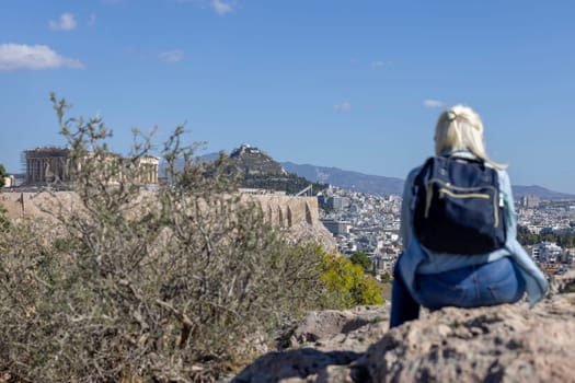 Athens, Greece - October 17, 2022:  Blonde woman sitting on a stone on Mouseion Hill and observing the beautiful view of Acropolis of Athens and Mount Lycabettus