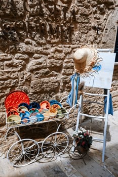 Alcudia Mallorca, Rustic Mediterranean decor: Colorful ceramic plates in vintage metal cart, straw hat on ladder, stone wall background. Traditional European street decoration