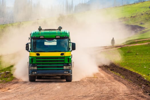 A truck kicks up dust on a dirt road with a pedestrian walking through the haze on a sunny spring day.