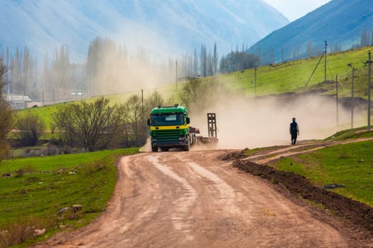 A truck kicks up dust on a dirt road with a pedestrian walking through the haze on a sunny spring day.