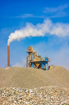 white steam rising up from pipe of small asphalt factory at day time with piles of gravel in a foreground.