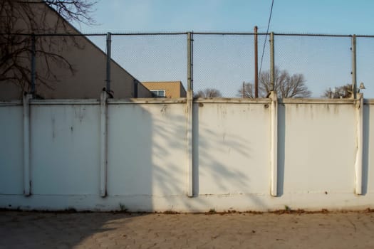 A weathered white wall topped with a chain-link fence, casting tree shadows across its surface in an open urban setting. The image captures themes of simplicity, urban architecture, and quiet solitude.