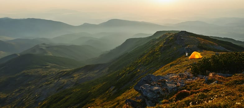 Man spending summer vacation in mountains, standing by tent looking at sunrise over valley. Active man hiking in mountains. Yellow tent put over valley. Foggy mountain landscape view at sunrise