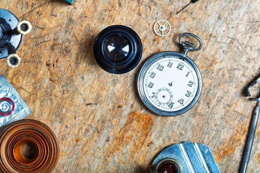 assortment of watchmaking tools surrounds a pocket watch on a worn wooden surface, displaying components like a gasket coil, magnifier, and gear, highlighting precision work