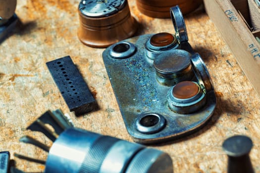 Watchmaker's workbench holds specialized tools, including a metal plate with containers, a perforated block, and a cylindrical tool, emphasizing the precision equipment needed for intricate repairs