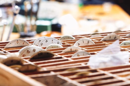 Wooden organizer filled with watch dials in individual compartments. Each dial displays unique markers, neatly arranged to prevent damage. The lighting and blur emphasize order in watch craftsmanship