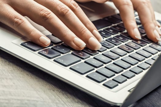 Hand with light skin and polished nails typing on a laptop, with a soft-toned background and coffee cup, adding focus to a peaceful workspace