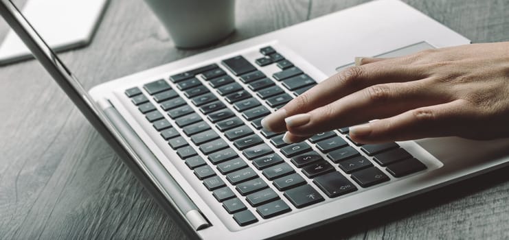 Light skin and well-manicured nails on a hand pressing a laptop key. The background shows a coffee cup, adding to the focused, calm workspace ambiance