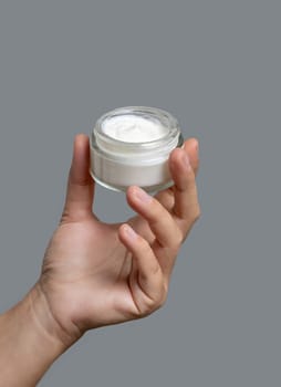 Female hand holding cream jar against grey background close up. Natural product for facial care, everyday skincare routine. Opened cosmetic Container. Spa and Wellness concept