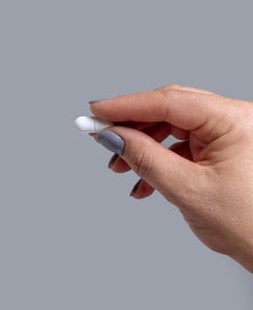 Female hand holding a white pill with fingers closeup against grey background. Healthcare, medical and pharmaceutical concept.