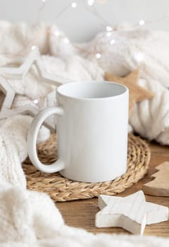 White blank coffee mug near white knitted sweater, wooden Christmas decor and lights, copy space. Winter atmospheric mock up in rustic or scandinavian style