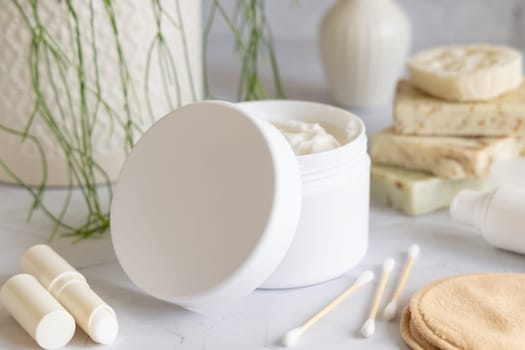 Opened cream jar with white blank lid on white marble against potted green plant and soap bars in bathroom, close up, packaging mockup. Cosmetic product for bodycare, beauty routine