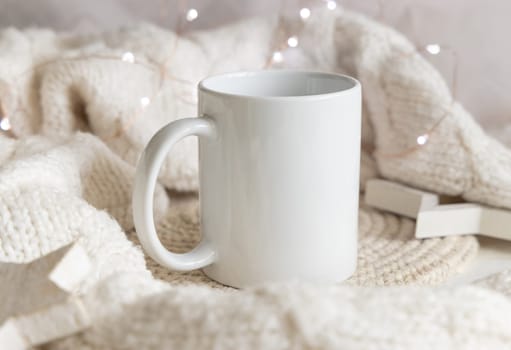 White blank coffee mug near white knitted sweater, wooden Christmas decor and lights, copy space. Winter atmospheric mock up in rustic or scandinavian style