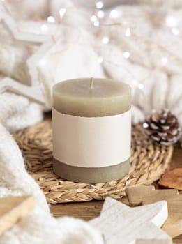 Pillar candle with blank label near white knitted sweater, wooden Christmas decor and lights, copy space. Winter atmospheric mock up in rustic or scandinavian style