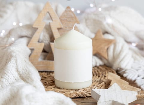 Pillar candle with blank label near white knitted sweater, wooden Christmas decor and lights, copy space. Winter atmospheric mock up in rustic or scandinavian style