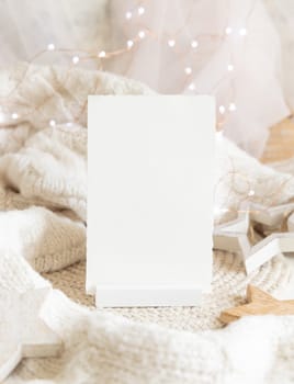 Vertical blank paper card near white knitted sweater, wooden Christmas decor and lights, copy space. Winter atmospheric mock up, family greeting in rustic or scandinavian style