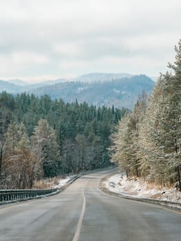 Winter road in snowy mountains. Autumn or wintertime in mountains. Asphalted road through fir tree forest and white snowy mountain peaks in the distance on background. Amazing landscape. Copy space