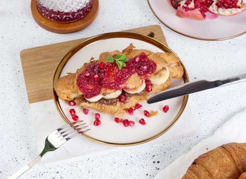 Croissant with banana pieces, chocolate paste and pomegranate seeds in a round white plate
