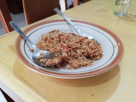 A plate of flavorful fried rice is served with a spoon and a fork on a wooden table. The dish includes pieces of vegetables and meats, showcasing a homemade and appetizing meal.