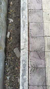 An urban scene showing the contrast between a patterned sidewalk and a dirt pathway. The textured surfaces highlight urban decay and natural elements coexisting in a city environment.
