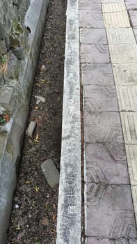 An urban scene showing the contrast between a patterned sidewalk and a dirt pathway. The textured surfaces highlight urban decay and natural elements coexisting in a city environment.