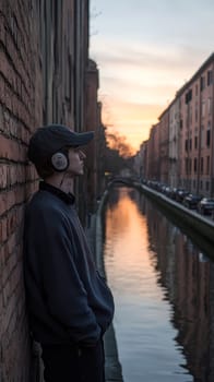 A young man leaning against a wall near the canals, wearing an oversized sweater and a baseball cap, listening to music through his vintage headphones. AI generative.