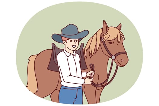 Cowboy boy is standing next to horse caring for stallion and wanting to become professional jockey and race. Schoolboy wearing texas ranger hat near horse for ride or competition at racetrack