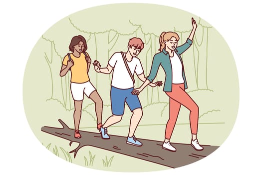 Group of friends participate in hike balancing on log thrown across river instead of bridge. Guy and two girls enjoy outdoor activities and hike in nature reserve with impenetrable terrain