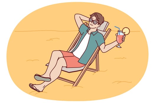Man tourist is sunbathing on beach sitting in sun lounger and drinking fruity refreshing cocktail. Guy tourist and travel lover sunbathes spending vacation on beach of sunny ocean.