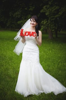A joyful bride, dressed in a stunning dress, holds a sign of love in her hands, surrounded by lush, bright greenery
