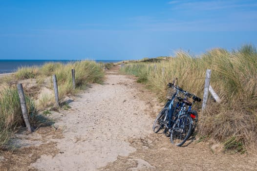 Footpath over the sand dunes on a beach by the sea, with parked bicycles at the fence, with blue sky