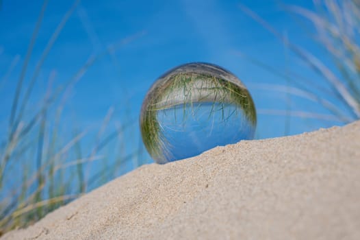 A glass ball lies in the sand of a dune by the sea, the dune grass and the blue sky are reflected in the ball