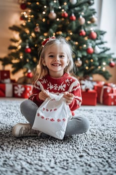 A joyful child in a cozy sweater sits on a rug, excitedly opening a gift beside a beautifully decorated Christmas tree.