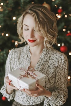 A happy woman with blonde hair smiles as she holds a beautifully wrapped gift near a decorated Christmas tree.
