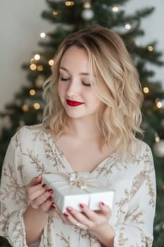 A woman with wavy hair holds a beautifully wrapped gift, smiling in a cozy room decorated for Christmas with shining lights.