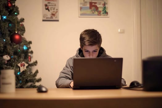 A young boy sits at a wooden table, intently working on a laptop, surrounded by holiday decorations and a Christmas tree.