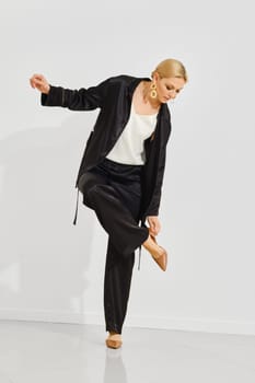 A woman stands gracefully indoors, wearing a sleek black outfit and adjusting her shoe.