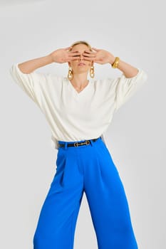 A model stands confidently in a minimalist studio, striking a pose while blocking her face with her hands. She wears a white top and vibrant blue pants, emphasizing fashion and modern style.