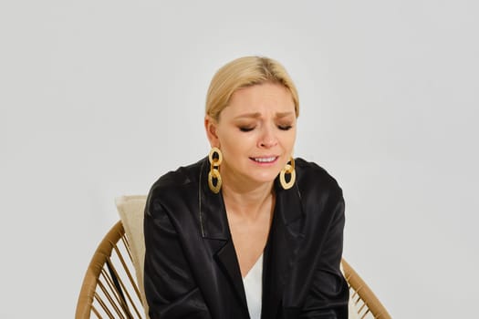 A woman with blonde hair sits in a rattan chair, visibly emotional as she reflects. She wears large earrings and a black jacket, creating a contrast with the minimalistic backdrop.