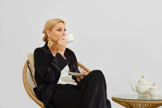 A woman is peacefully sipping tea from a delicate cup while sitting comfortably on a rattan chair. She is dressed in a casual outfit, creating a relaxed atmosphere in a minimalist room.