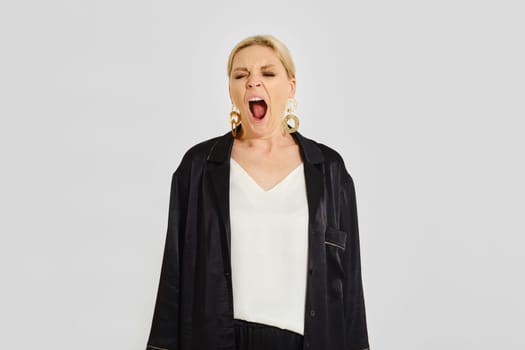 A woman stands in a minimalist studio and yawns, showcasing her relaxed demeanor. She wears a simple top and black outerwear, accessorized with bold earrings, highlighting a casual yet stylish look.