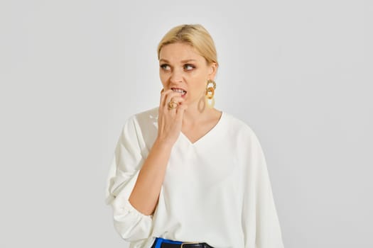 A woman with blonde hair styled elegantly wears a loose white blouse and large earrings, displaying a nervous gesture as she bites her nails against a neutral background.