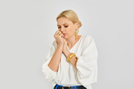 In a minimalist setting, a woman with blonde hair stands with her arms crossed, showing a moment of contemplation and vulnerability. She wears a white top and is adorned with jewelry.