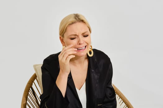 Crying woman with blonde hair sits in a stylish chair, tearing up and expressing deep emotions.