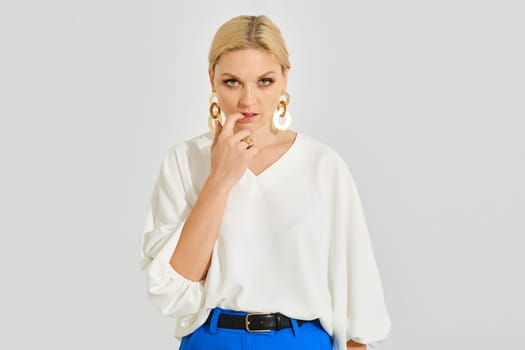 The woman stands confidently with a contemplative expression, dressed in a white top and vibrant blue pants and bites her nail. Her striking accessories add elegance to her contemporary look.