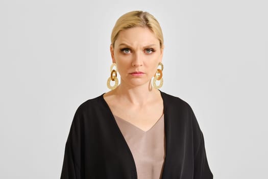 A woman stands tall with a serious expression, wearing large gold earrings and a dark outfit. Her blond hair is styled elegantly, creating a striking contrast with the simple background.
