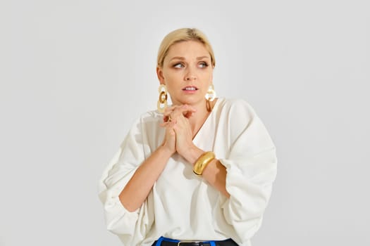 A woman with blonde hair, wearing a white blouse and statement earrings, displays a nervous expression. The backdrop is simple and unadorned, contributing to a focused ambiance.
